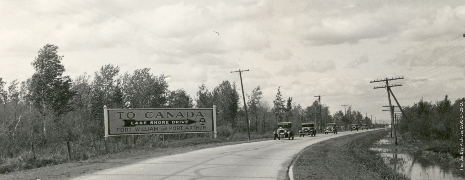 black and white photo of a road sign to Port Arthur and Fort William and old cars on driving on road