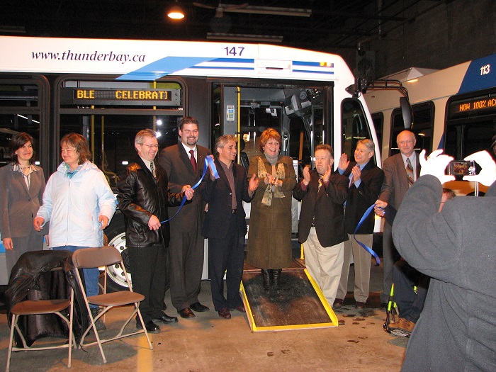 A group of people stand in front of a bus with a ramp and a ribbon is cut
