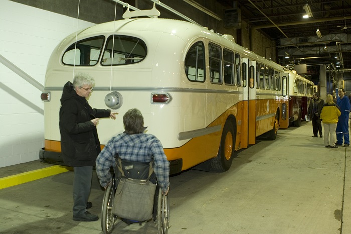 A man in a wheelchair being shown a pre-1970s orange and white bus