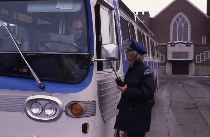 a man standing beside a bus driver's side window talking to a bus driver inside of the bus.
