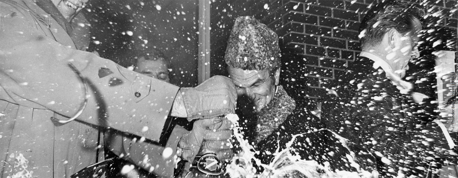 Four men smashing a bottle of champagne as it sprays