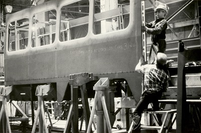 Workers standing on ladders as they place pieces on a partially assembled bus