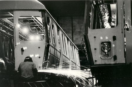 a welder working on the shell of a bus on his left, with another waiting to the right