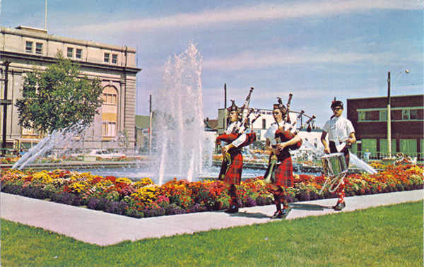 Two bagpipers playing in front of a water fountain with a building in back