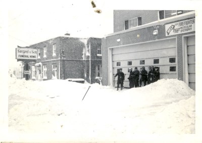 A group of firefighters standing outside a fire station with shovels in the snow