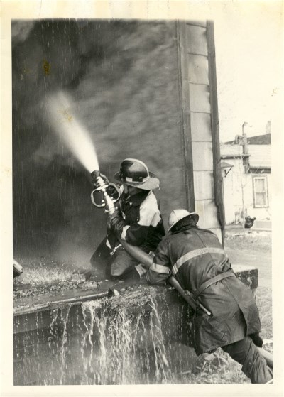 Two fireman holding a hose and putting out a fire while standing on a ladder 