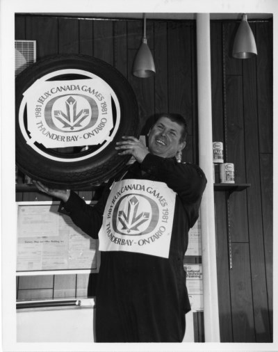A man stands infront of, and holding a Canada Games branded tire