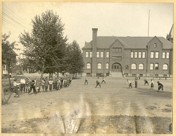 A field with a building in the background and children playing baseball out front