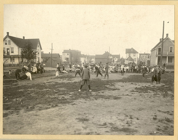 Boys playing a baseball game with people watching and houses in background