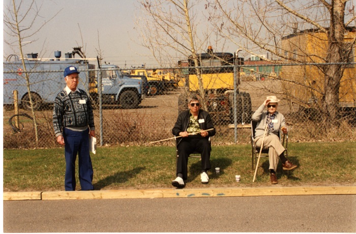 Two men sitting holding measuring sticks with a third man standing to the left