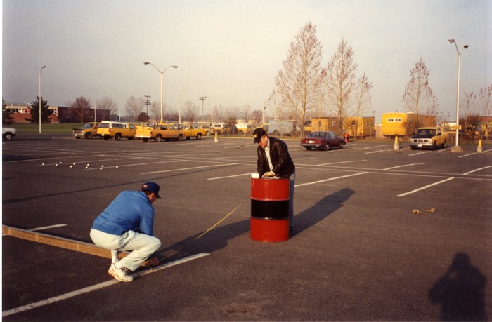 Two men measuring the distance between a barrel and a white line with a ruler