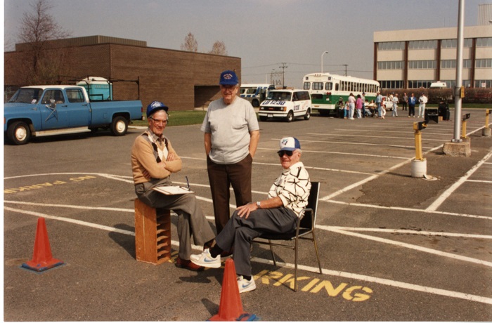 Two men sitting while one man stands in a parking lot