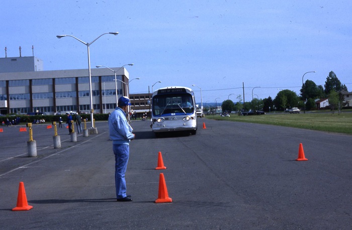 A man standing waiting for a bus to drive down a lane with orange cones on either side
