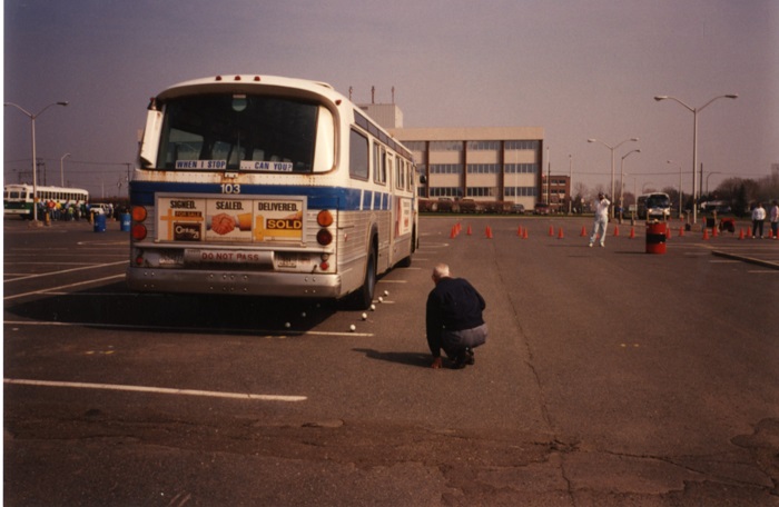 A man squatting on the ground looking to see where the wheels of a bus are