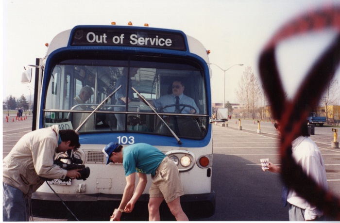 A man videotaping another man measuring something in front of a bus