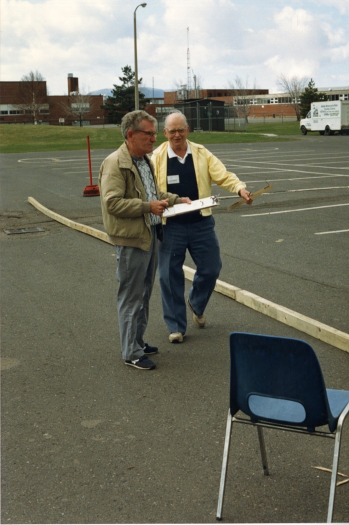 Two men standing talking and looking at a clipboard in a parking lot