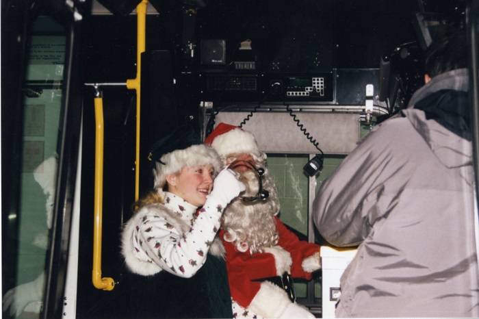 Santa driving a city bus with a helper jingling bells beside him