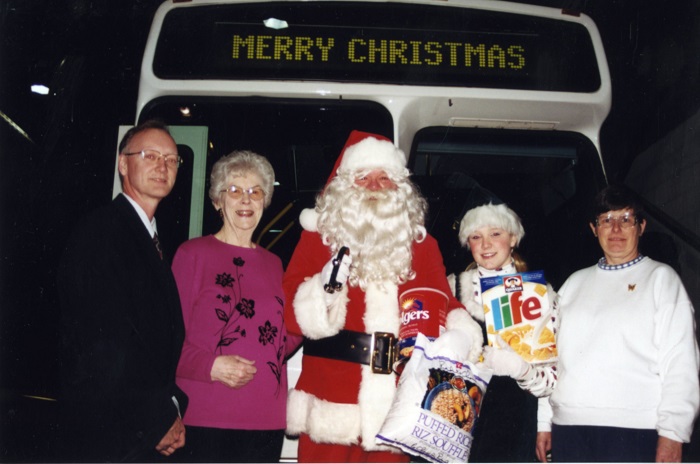 Santa in front of a bus with his helper holding donated food items.