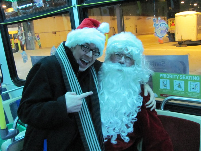 Santa sitting on a bus with a person sitting on his lap and pointing towards him