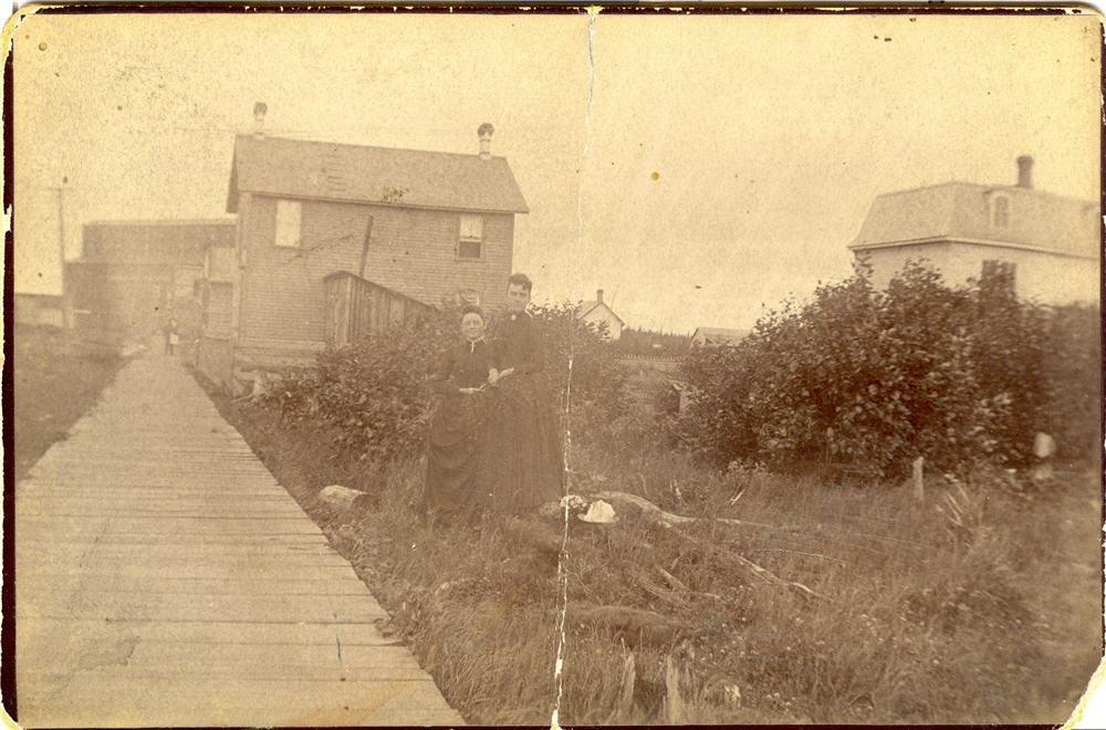 Two women stand together in front of severeal houses beside a wooden boardwalk