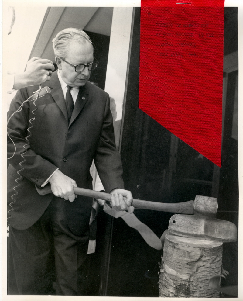 A black and white photo of a man cutting a ribbon with an axe - a piece of red ribbon is attached