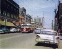 A trolley going down the street with cars parked on either side