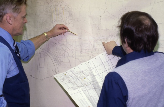 Two people looking at a map hanging on a wall, both are pointing at different places
