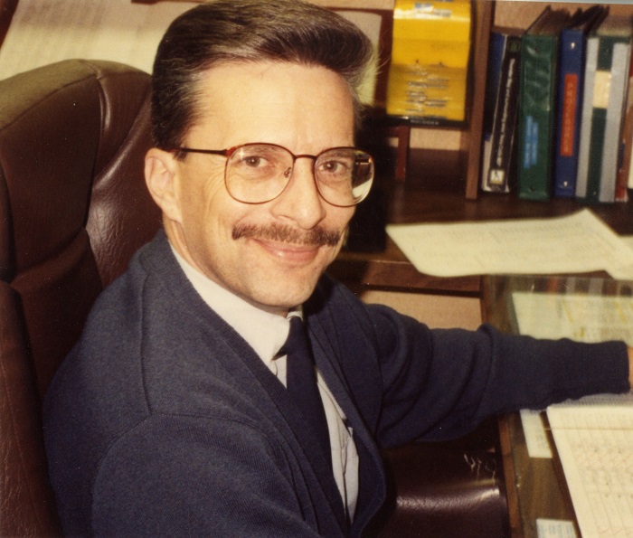 A man sitting and turning towards the camera smiling, he has large glasses