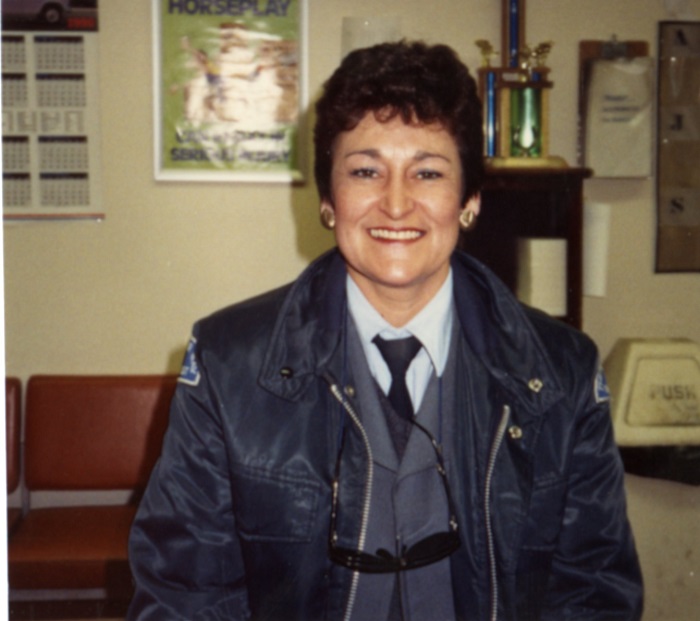 A woman wearing a jacket who has short hair standing and smiling at the camera