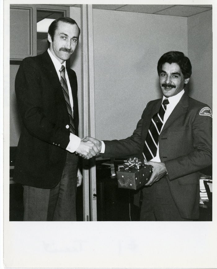 Two men stand shaking hands, one holding a gift in his other hand