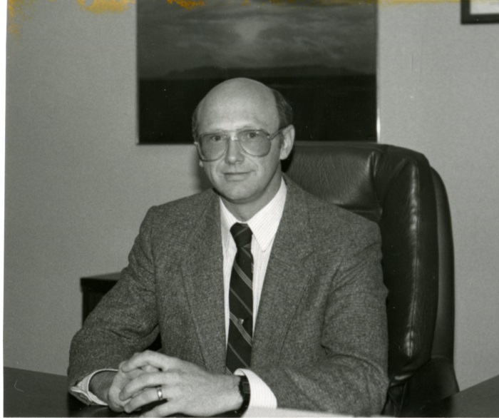 A man wearing glasses sitting behind a desk in a large chair