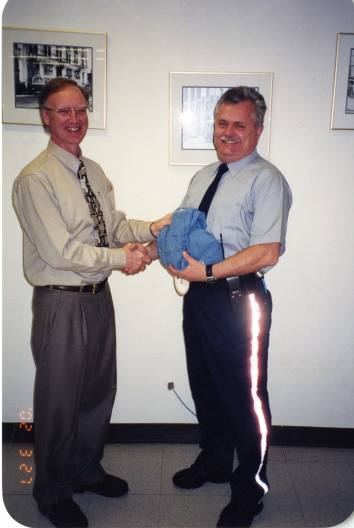 Two men standing and looking at the camera while shaking hands, one holds a shirt