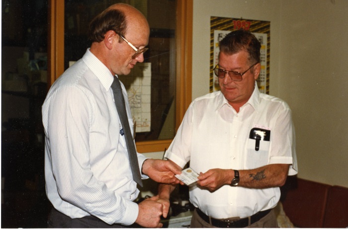 two individuals, one handing the other a card and they are shaking hands