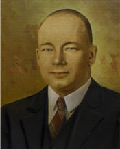Mayor Cowan wears sits wearing a black suit and brown tie