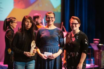 two women presenting a third individual with an award with band in background on stage