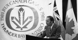 A man standing at a podium wearing a suit with flags and a banner behind