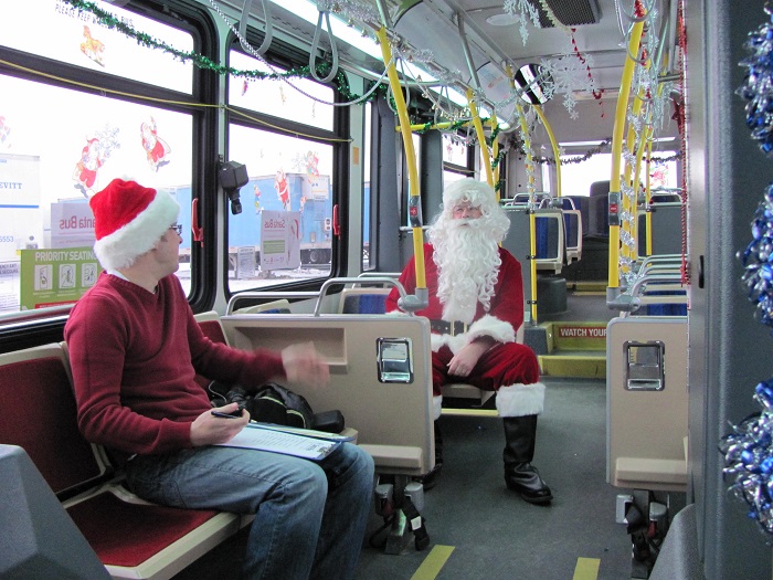 Santa sitting on a bus with a man wearing a snata hat seated a few rows ahead