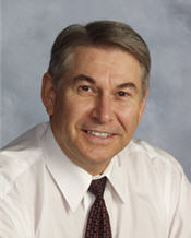 Mayor Ken Boshcoff sitting in front of a white and grey background wearing a shirt and tie