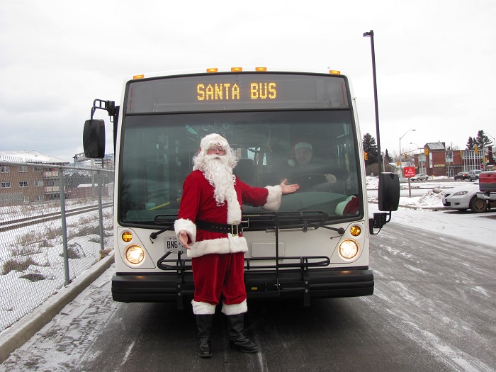 Santa standing in front of a bus with a 'Santa Bus' sign gesturing at the driver