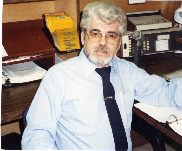 A man wearing a buttoned shirt, tie and glasses sits and smiles at the camera in an office