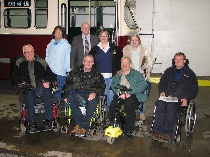 A group of people both standing and seated in wheelchairs pose in front of a trolley