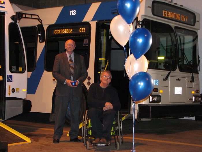 One man standing, while another is seated in a wheelchair in front of a bus