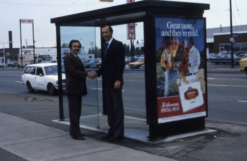 two men shaking hands in front of bus shelter.