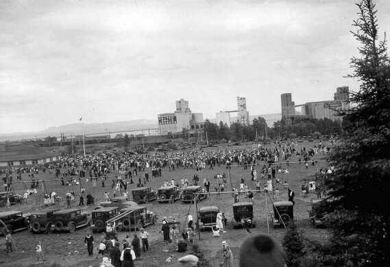 Dozens of cars sitting in a park with swing sets and people playing