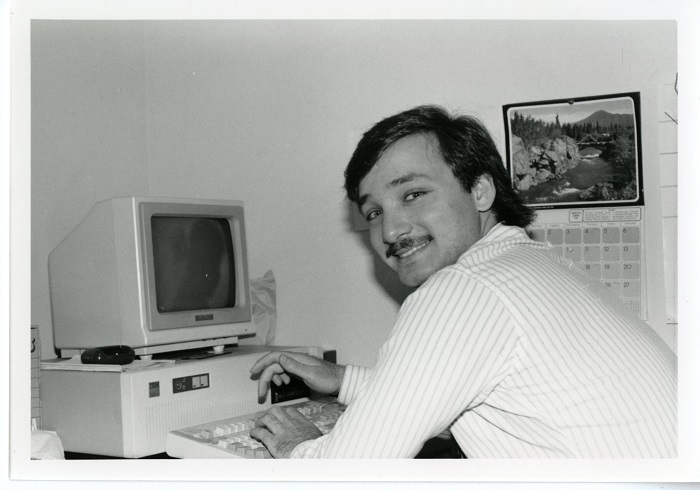 A man turning in his seat and smiling while seated at a desk with a computer