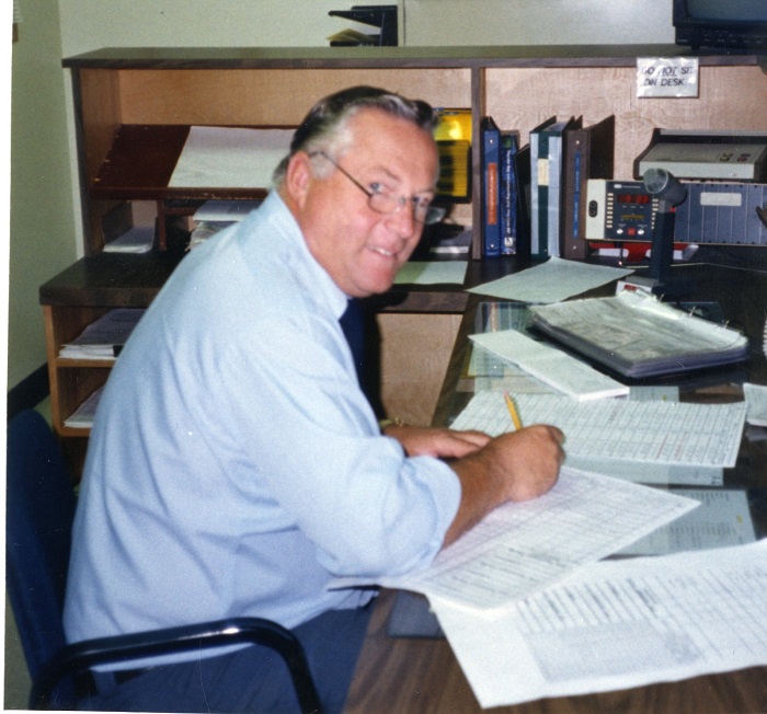 A man sitting at the desk writing while he turns to the camera and smiles