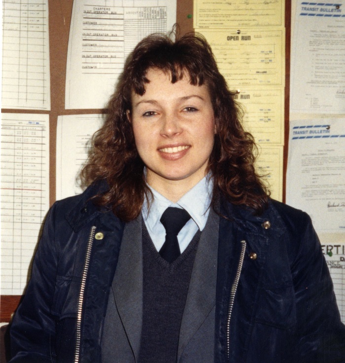 A woman standing and looking at the camera, wearing a jacket and has curly hair